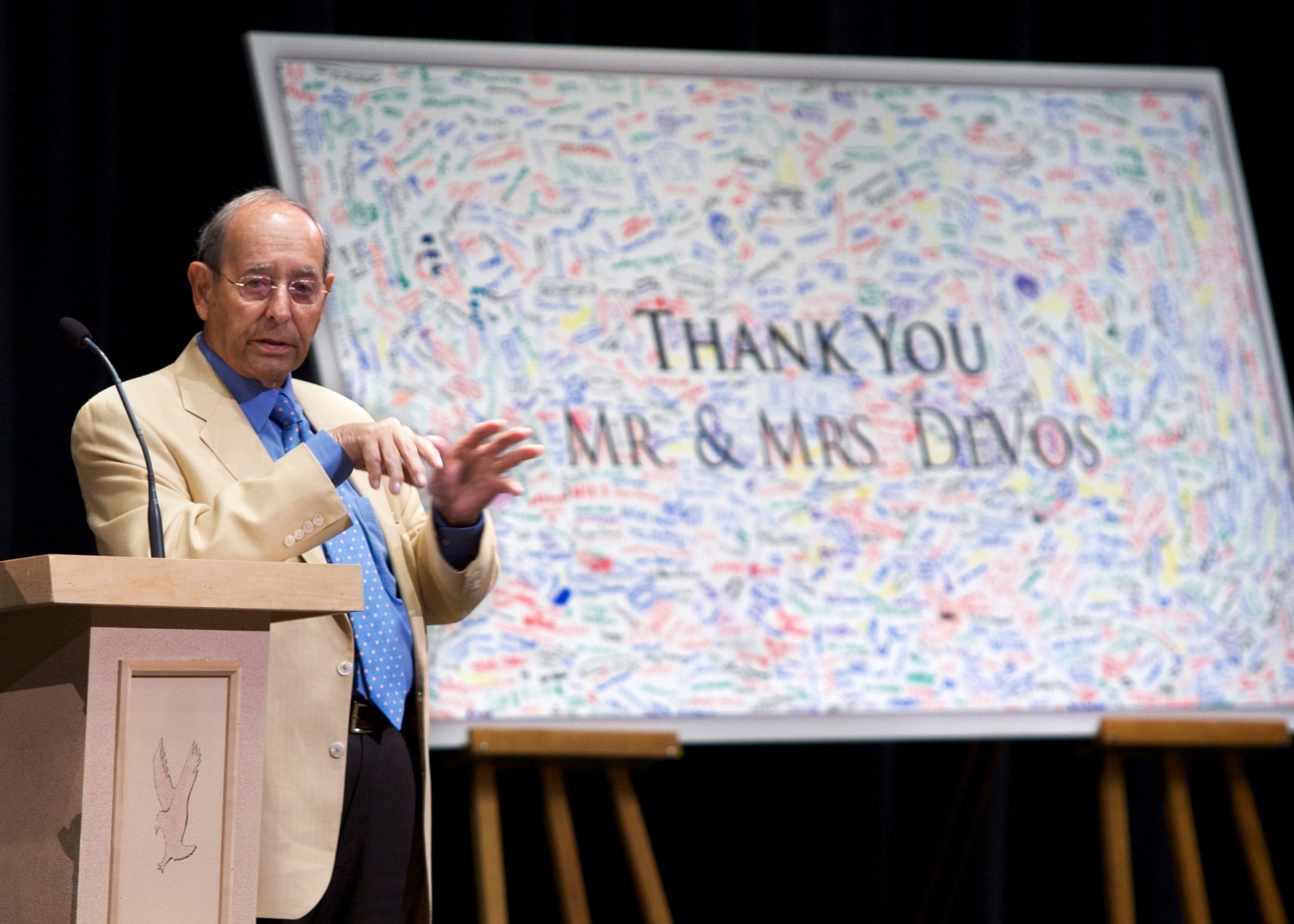 Rich stands near a podium while speaking to a crowd. A large white board reading "Thank you Mr. & Mrs. DeVos" is seen in the background.